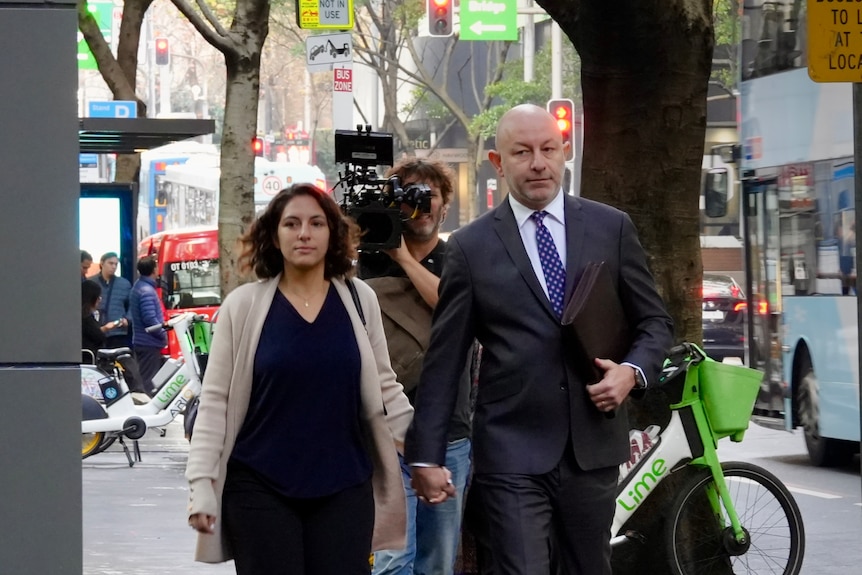 Mr Ostrovsky in a black suit and blue tie walking next to a woman in a black dress with a news camerman behind them.