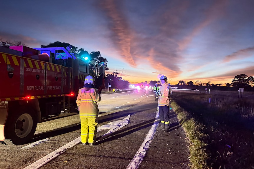 Two rfs members standing on highway next to truck in high viz and helmets, sky is lit with sunrise and colours.