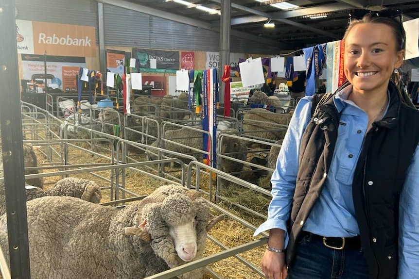 A woman stands smiling next to a sheep in a pen inside.