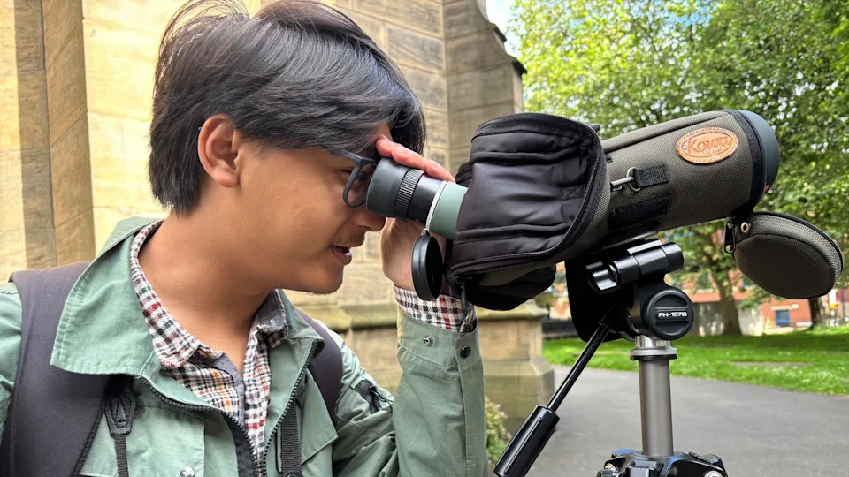 An man with dark floppy hair peers through the lens of a covered device like a telescope