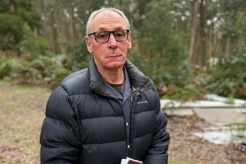 A man with white hair, dark rimmed glasses in a dark puffer jacket stands near trees holding a notebook in his clasped hands.