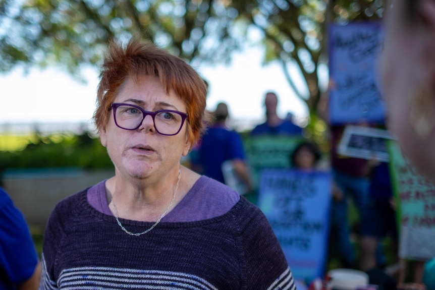 A woman in a purpole jumper and glasses speaking to left of frame with protesters in the background.