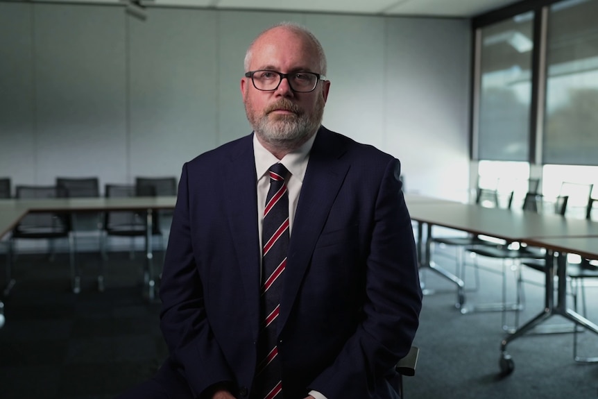 A balding man wearing a navy suit, white shirt, red and navy tie and glasses sits in a nondescript meeting room.