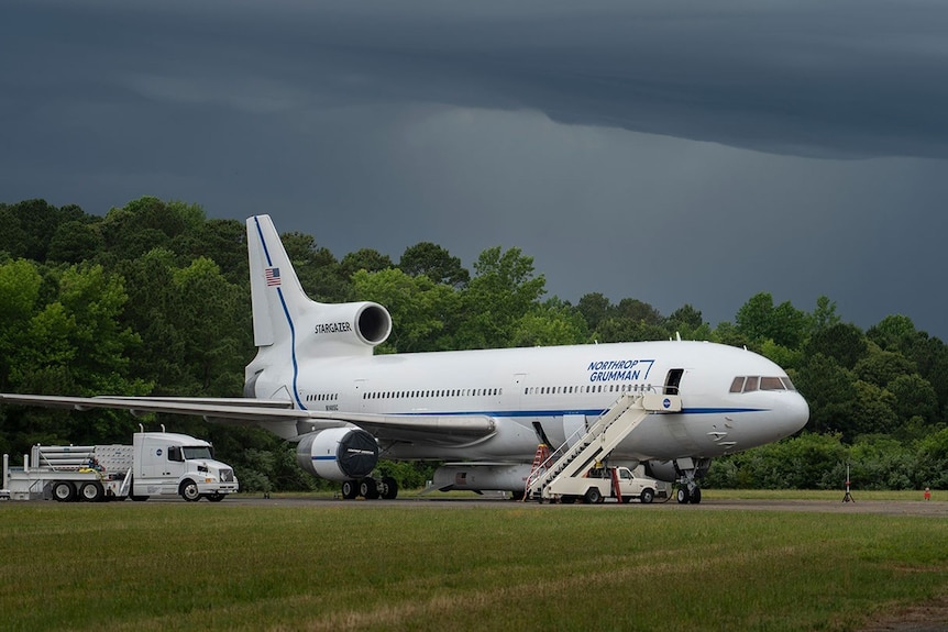 A white plane on a tarmac next to a truck and car, with blue 'Northrop Grumman' livery.