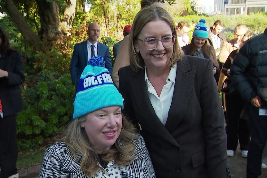 A woman in a blue beanie and checked blazer sits beside a woman with chin length light hair in a dark blazer who is standing.
