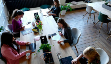 A group of women sit at a long table working on laptops