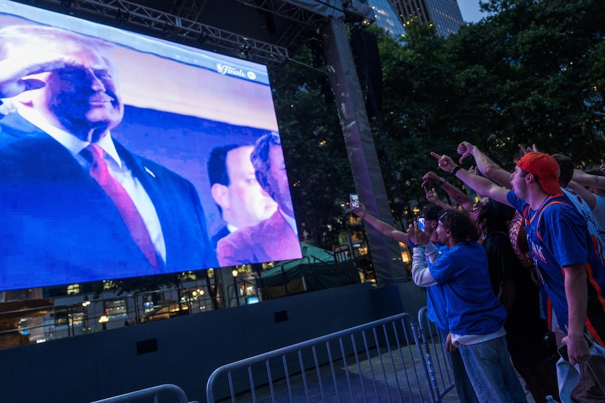 People watch a big screen which has Donald Trump making a salute.