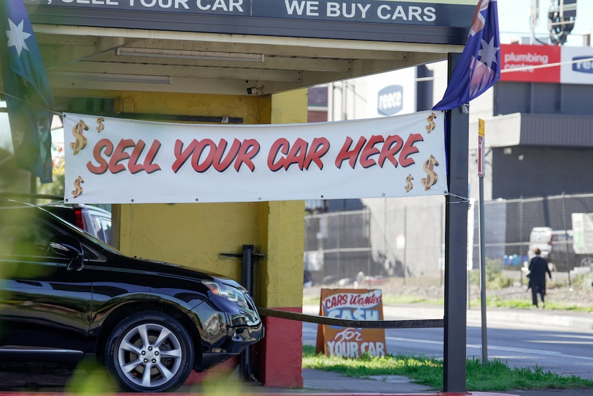 A generic car yard wide shot, with a black car parked outside a yellow building with a sign saying "Sell your car here".