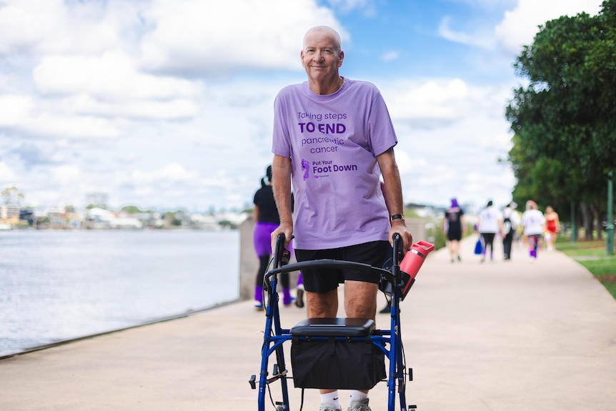 A man in a purple shirt stands behind a walker on a footpath alongside a river