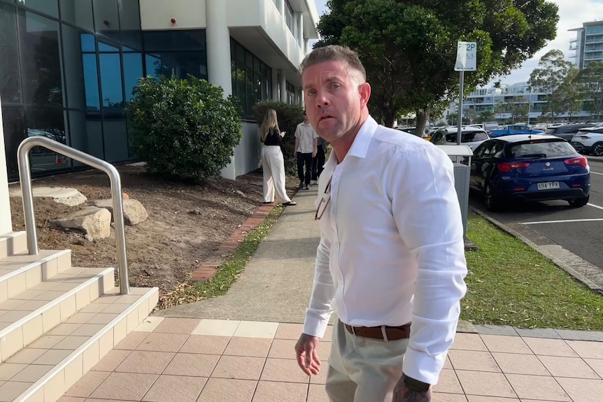 A white man with close-cropped hair, in a white shirt and light pants, walking towards steps.