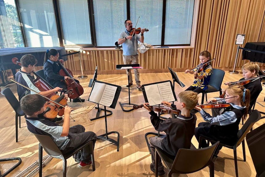 A group of young children sit in a circle with violins at their chins while an violin instructor stands before them