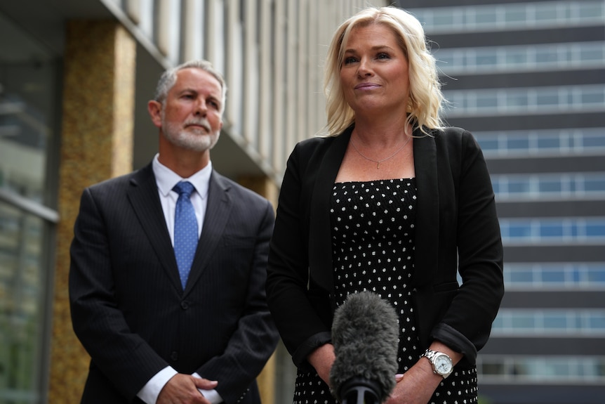 Leanne Castley stands in front of microphones, with Jeremy Hanson behind her looking at her.
