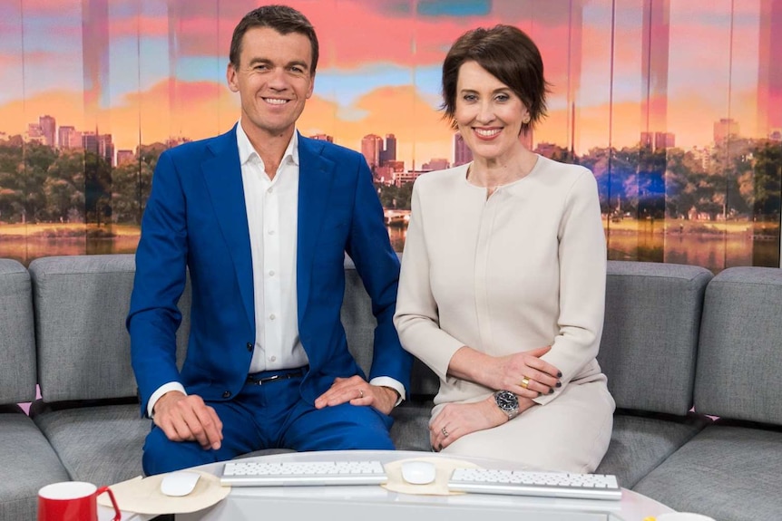 A man and a woman sitting on a couch on the set of a TV show, with a cityscape behind them.