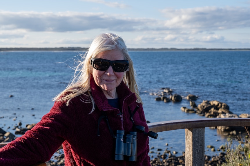 Older woman with shoulder length grey hair and burgundy jumper stands in front of ocean.