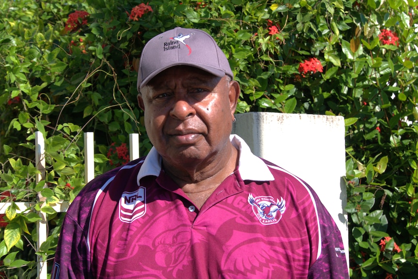 Falen with a serious face wearing a hat that says "Raine Island", a fuchsia pink and purple polo neck shirt with patterns.