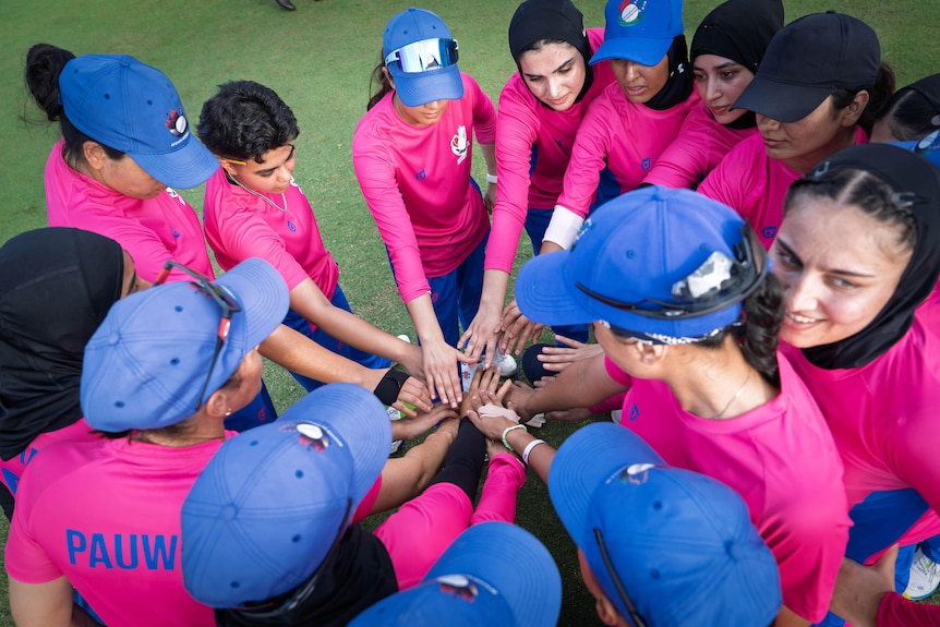 High-angle shot of huddle of Afghan women cricket players, hot-pink shirts, blue caps, standing on green sports field.