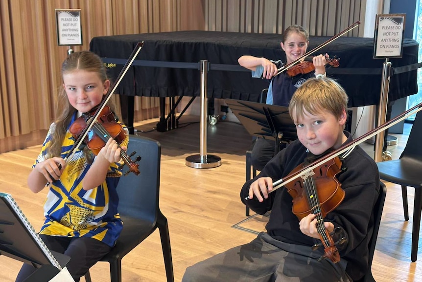 Three students hold violins below their chins and smile at the camera