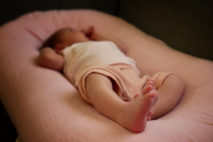 An infant lying on a pink pillow, wearing a white top, pink shorts, with little feet in focus and face out of focus