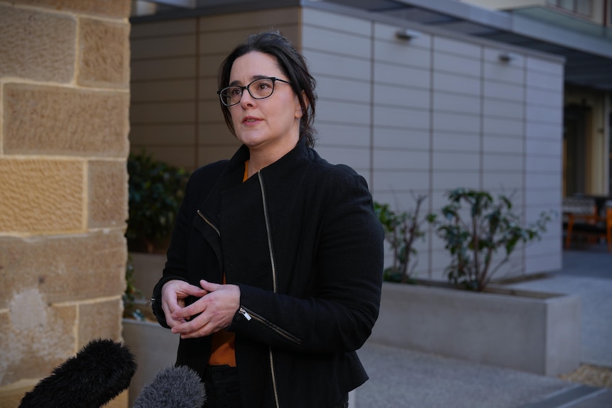 A woman with glasses and dark hair, wearing a dark jacket, speaks before microphones outside a brick building.