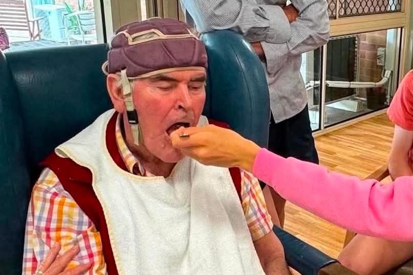 Elderly man in a helmet being fed by someone out of frame