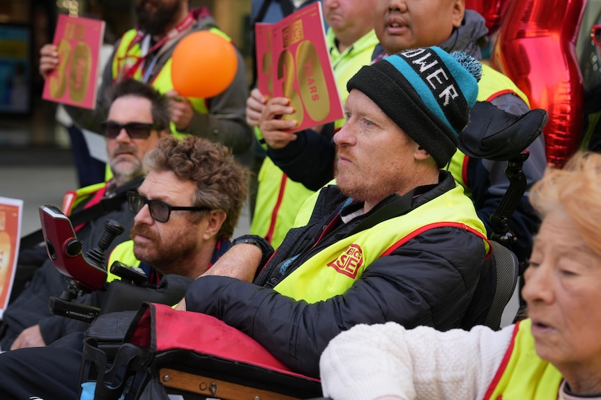 A man wearing a footy beanie and fluorescent yellow vest sits in a wheelchair among coworkers.