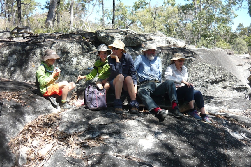 A family, two children, an adult woman, and an older couple sit on a large rock in the bush.