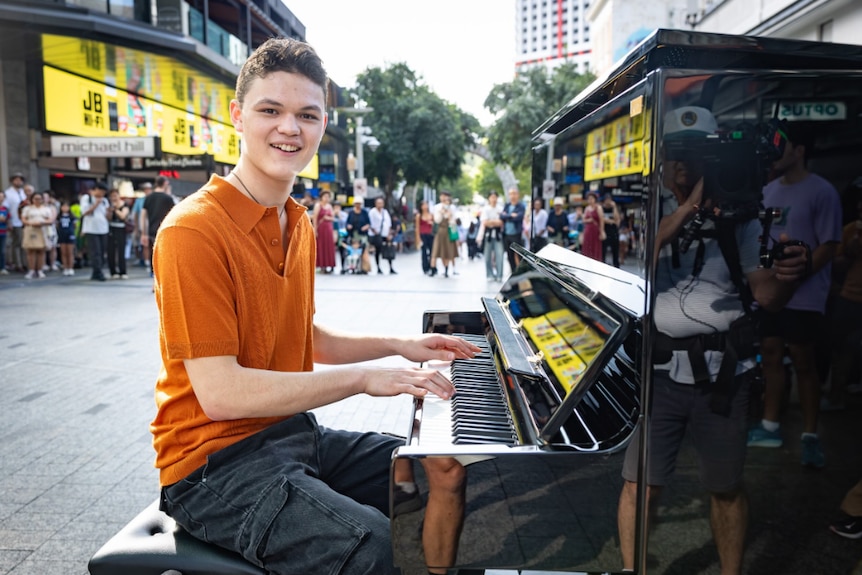 A smiling young man in an orange shirt is seated at a black upright piano in the middle of a busy pedestrian area.