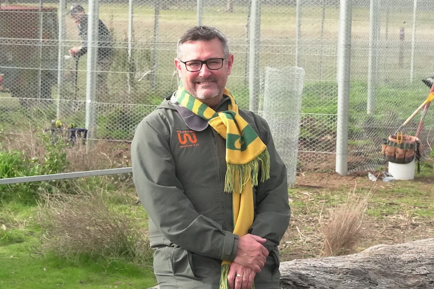A man with black and grey hair stands outside smiling near a fenced area, wearing a scarf.