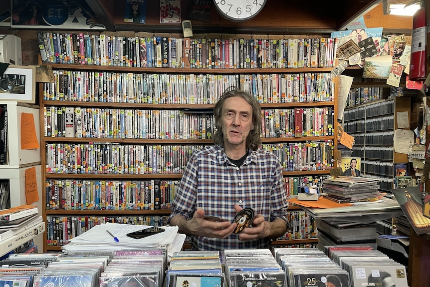 Man holds a DVD disc behind a shop counter with a CD display at the front and DVDs shelved along the wall.