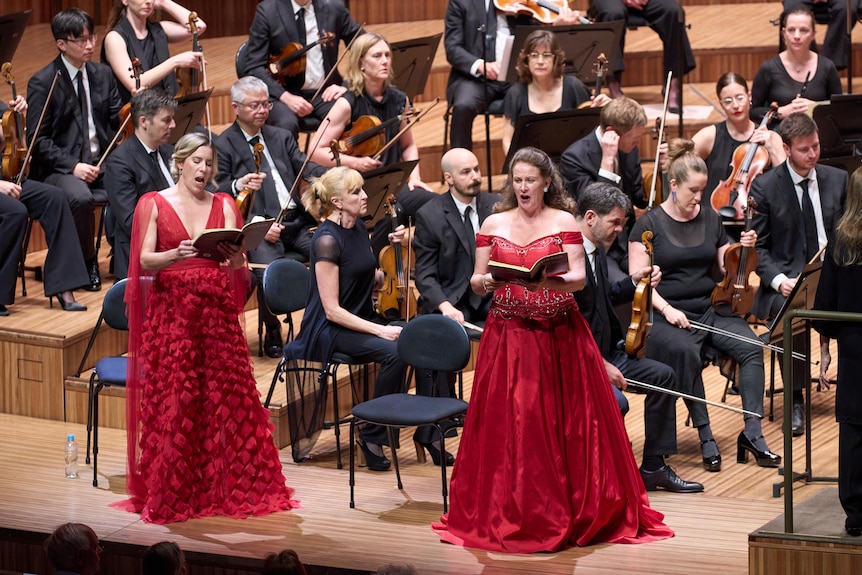 Two singers in red gowns perform to an audience, standing in front of a large orchestra.