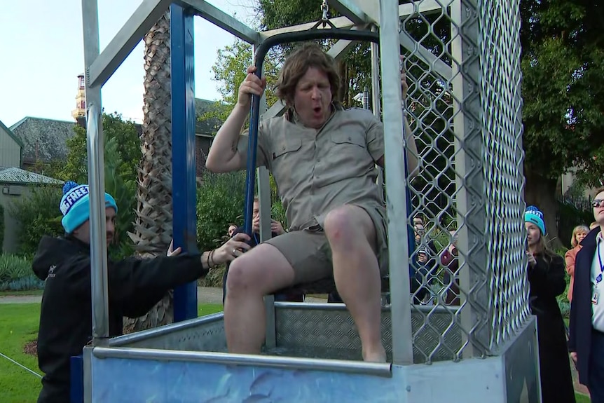 A man in a blonde wig and khaki shirt and shorts winces while sitting on a raised seat inside a steel cage.