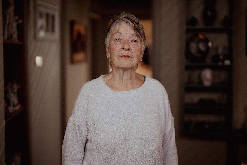 Older woman in a grey-purple jumper standing in a house.