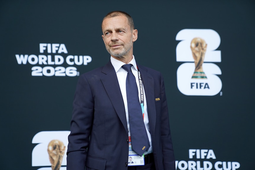 Aleksander smiles, looks sideways, wears blue suit, tie, Fifa World cup 2026 behind, Is Caucasian.