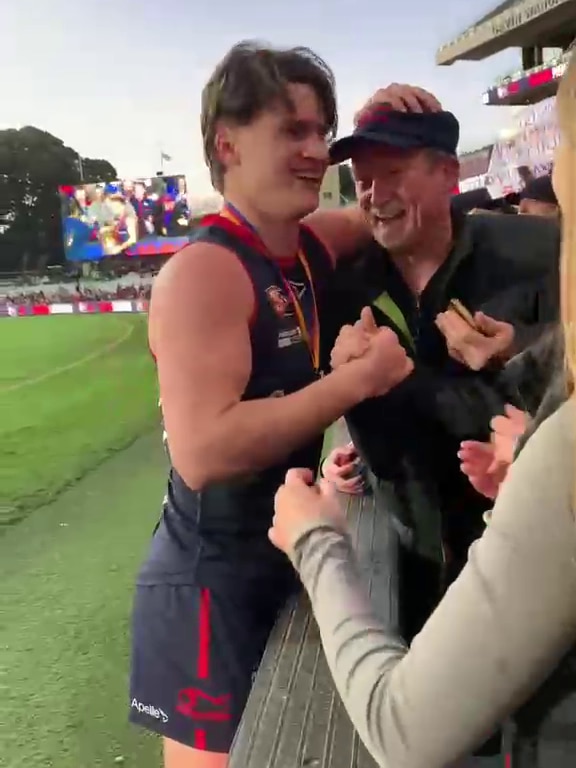 A young man in AFL gear and a man smile and put an arm around each other at the boundary fence of an AFL oval.
