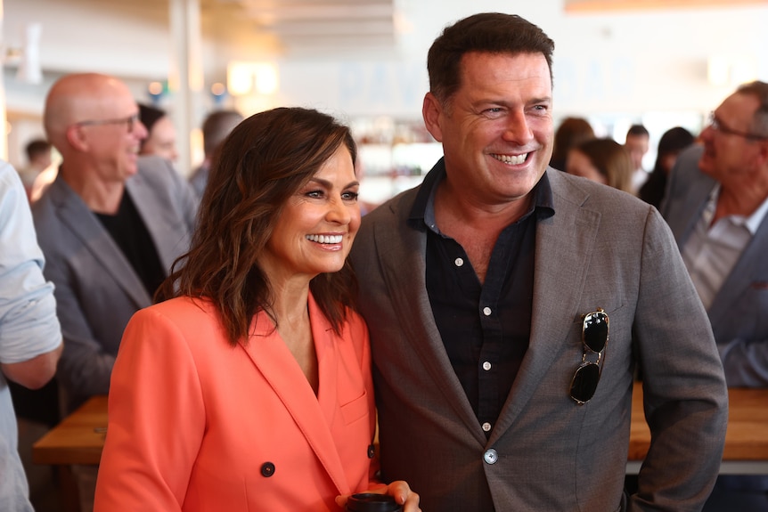 Lisa wears a bright suit and smiles next to Karl on a red carpet, who wears a dark suit.