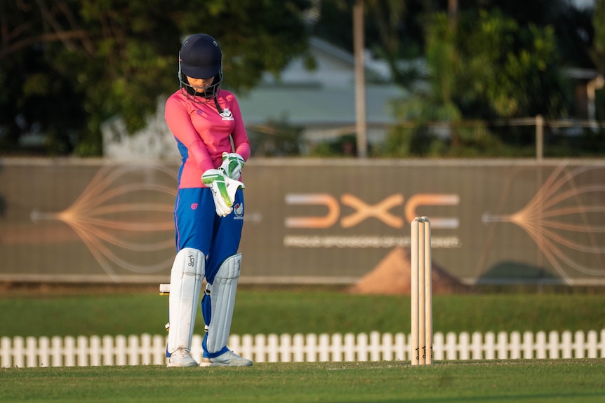 A sole women's cricket player, Afghan, hot-pink long-sleeve shirt, blue pants, knee pads and gloves on, standing on pitch.