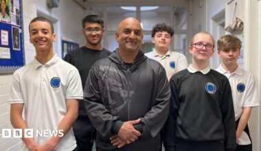 Aimran Majid stands in a classroom corridor surrounded by the students he teaches at Lindsworth School. He is wearing a black jacket and trousers, the five male and female students are wearing their school uniform.