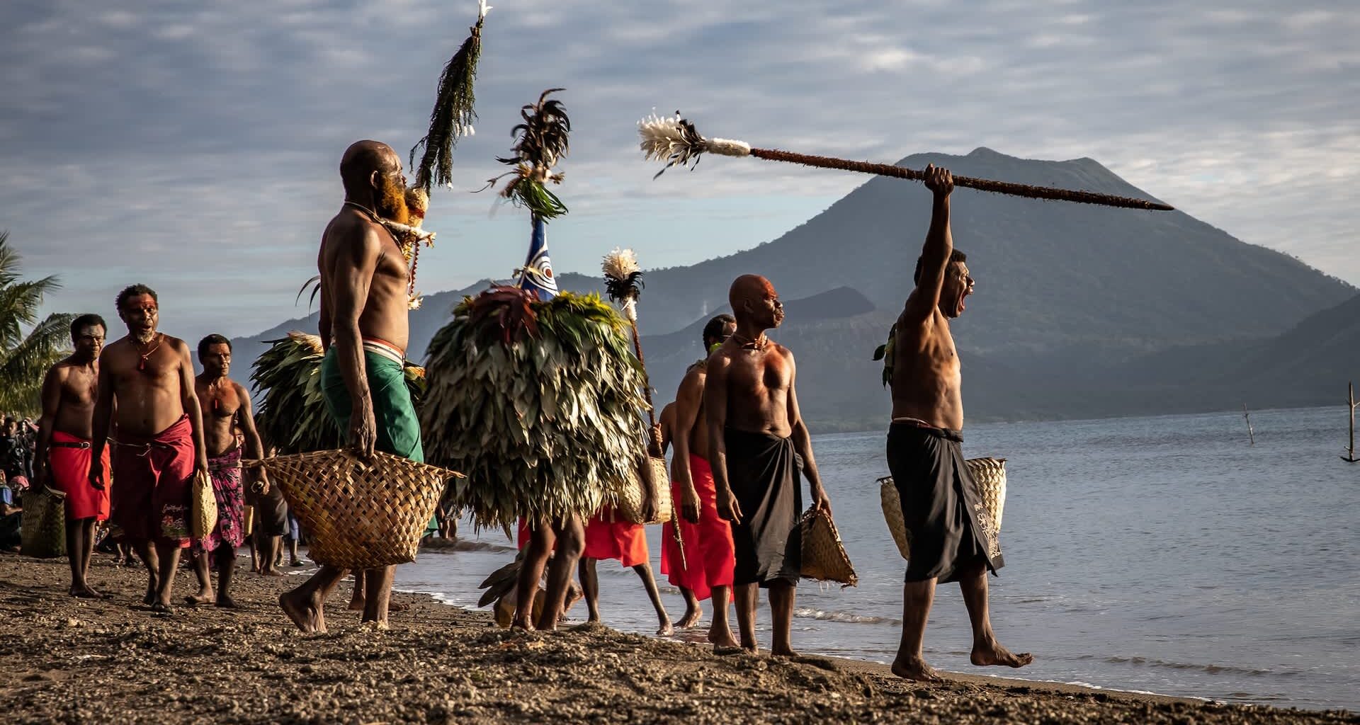 A group of men in traditional attire carry baskets and ceremonial objects along a beach with mountains in the background.