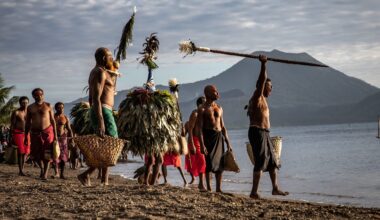 A group of men in traditional attire carry baskets and ceremonial objects along a beach with mountains in the background.