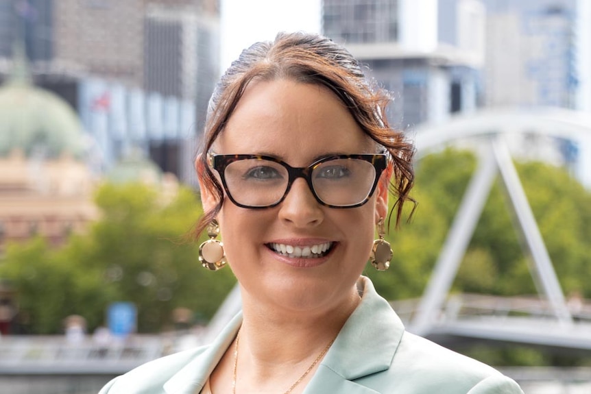 A woman with brown curly hair wearing a suit and a pair of glasses smiling at the camera.