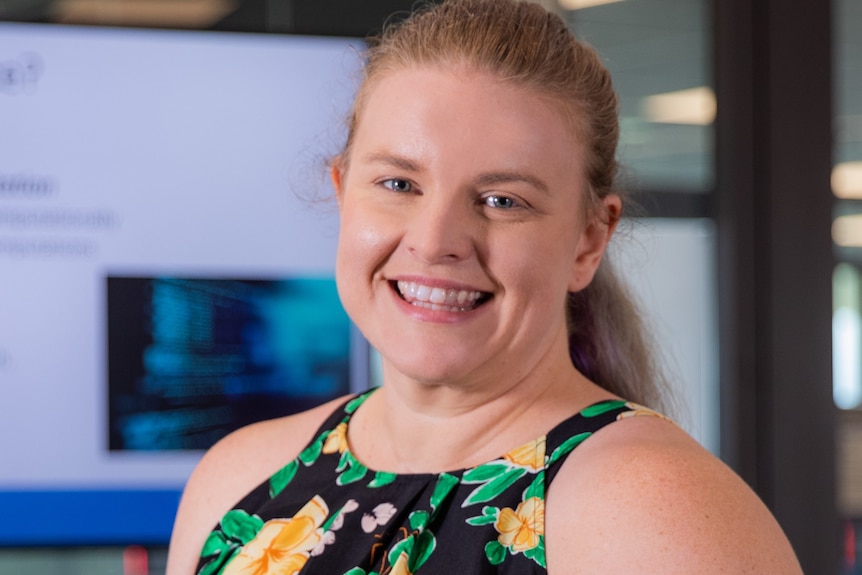 Blonde woman in floral blouse smiling standing in front of computer screens