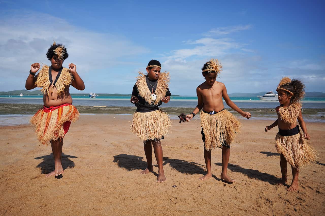 Four young Indigenous dancers on a beach. They are wearing traditional grass skirts, which are light brown, and woven grass headbands. Three of them have a grass 'garment' around their neck and covering their chests.