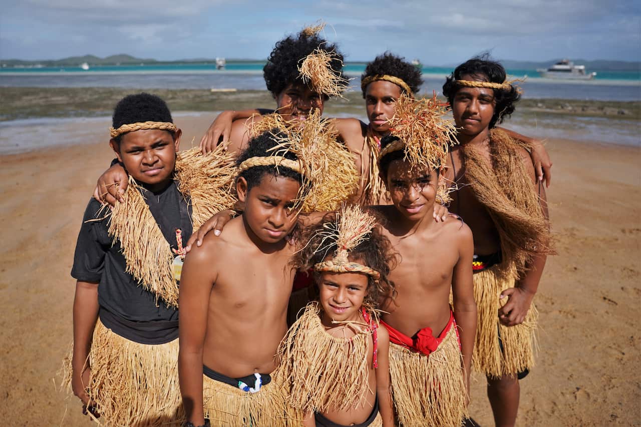 Seven young Indigenous people standing close together for a photo on a beach. They are wearing traditional grass skirts, which are light brown, and woven grass headbands. Four of them have a grass 'garment' around their neck and covering their chests.