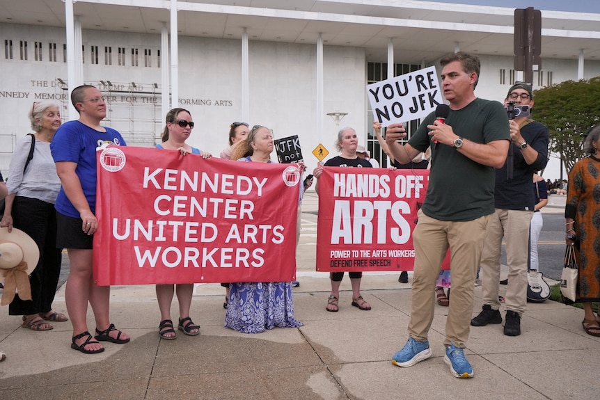 Men and women including former CNN anchor Jim Acosta speak outside the Kennedy Center, holding signs.