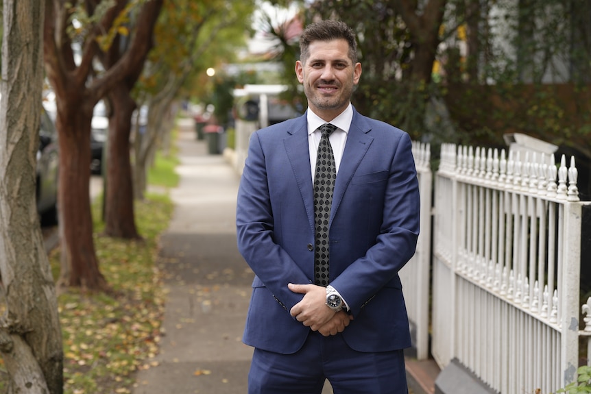 Michael wears a suit jacket and tie and stands in front of a residential street.