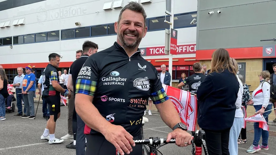 Image of a man in cycling gear. In the background can be seen a crowd of people and the outside of a large stadium building