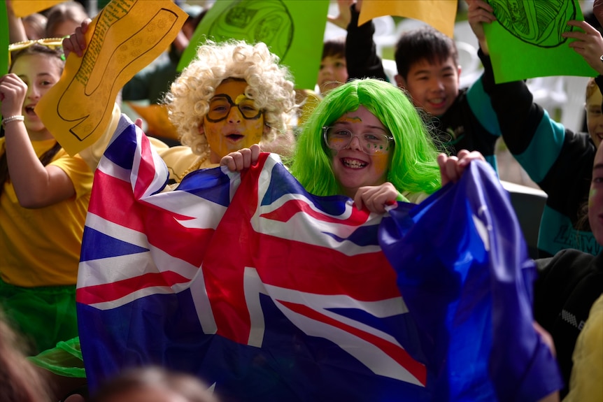 Two students, one wearing a gold curly wig and one wearing a bright green bob wig hold an Aussie flag.