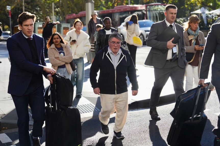 small grey-haired man in chinos and jumper crossing street surrounded by men in suits with suitcases 