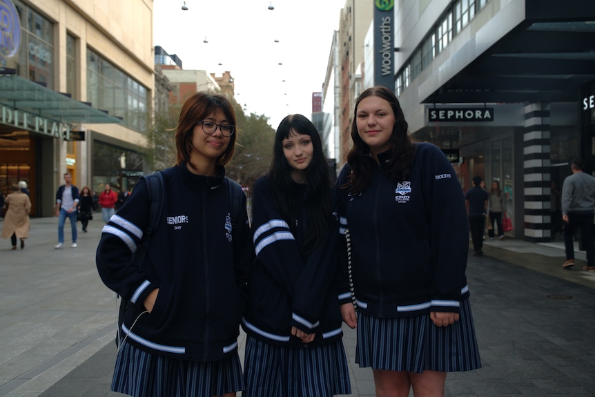 Three teenage girls wearing navy uniforms stand together in a shopping mall.