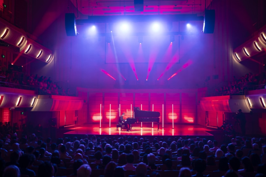 CJ Jones plays a grand piano on a brightly lit stage in a modern concert hall before a large seated audience